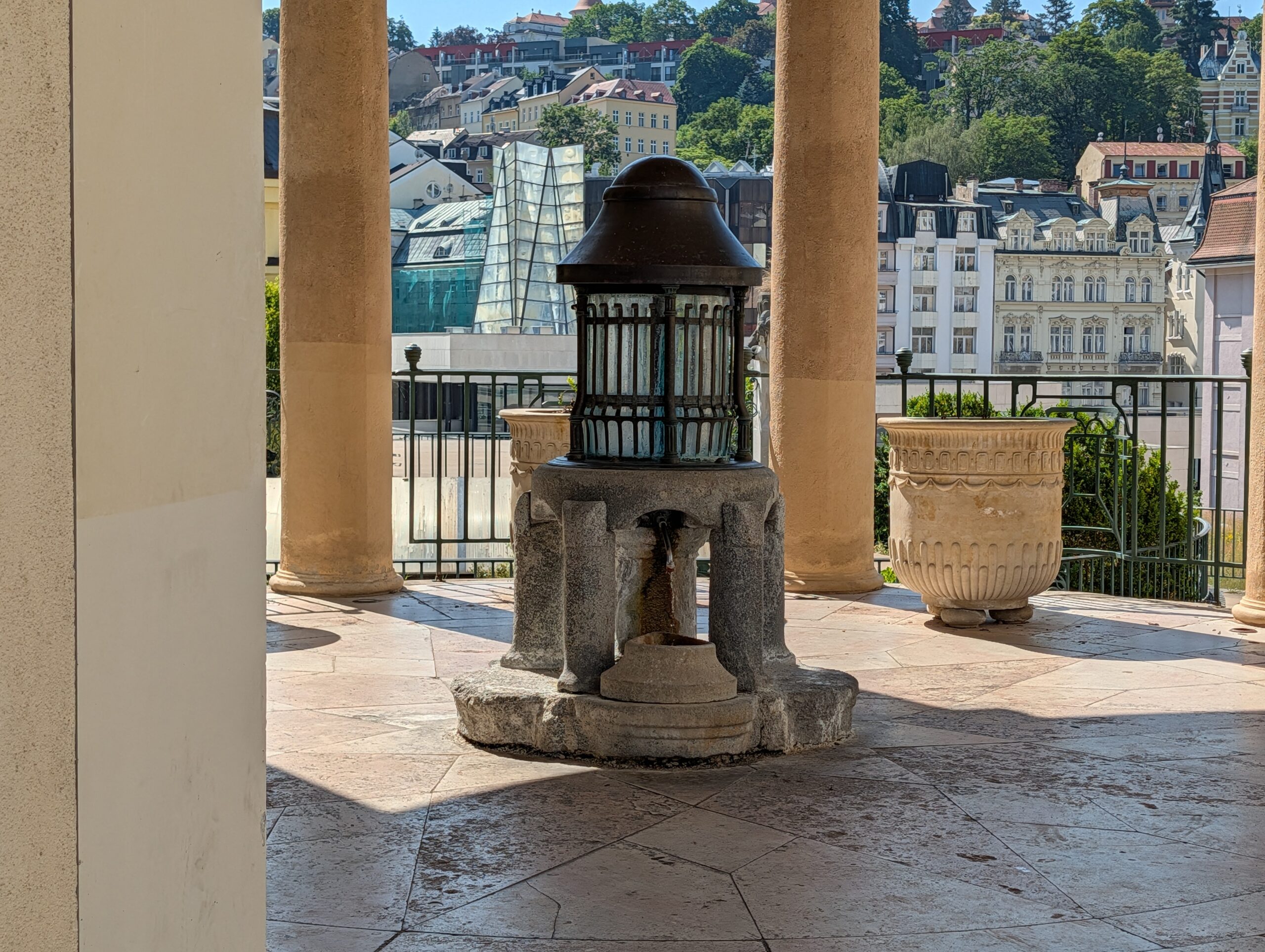 Oberer Schlossbrunnen Nr. 4 mit Metallgitter und Auffangbecken vor der Kulisse des Karlsbader Kurzentrums.