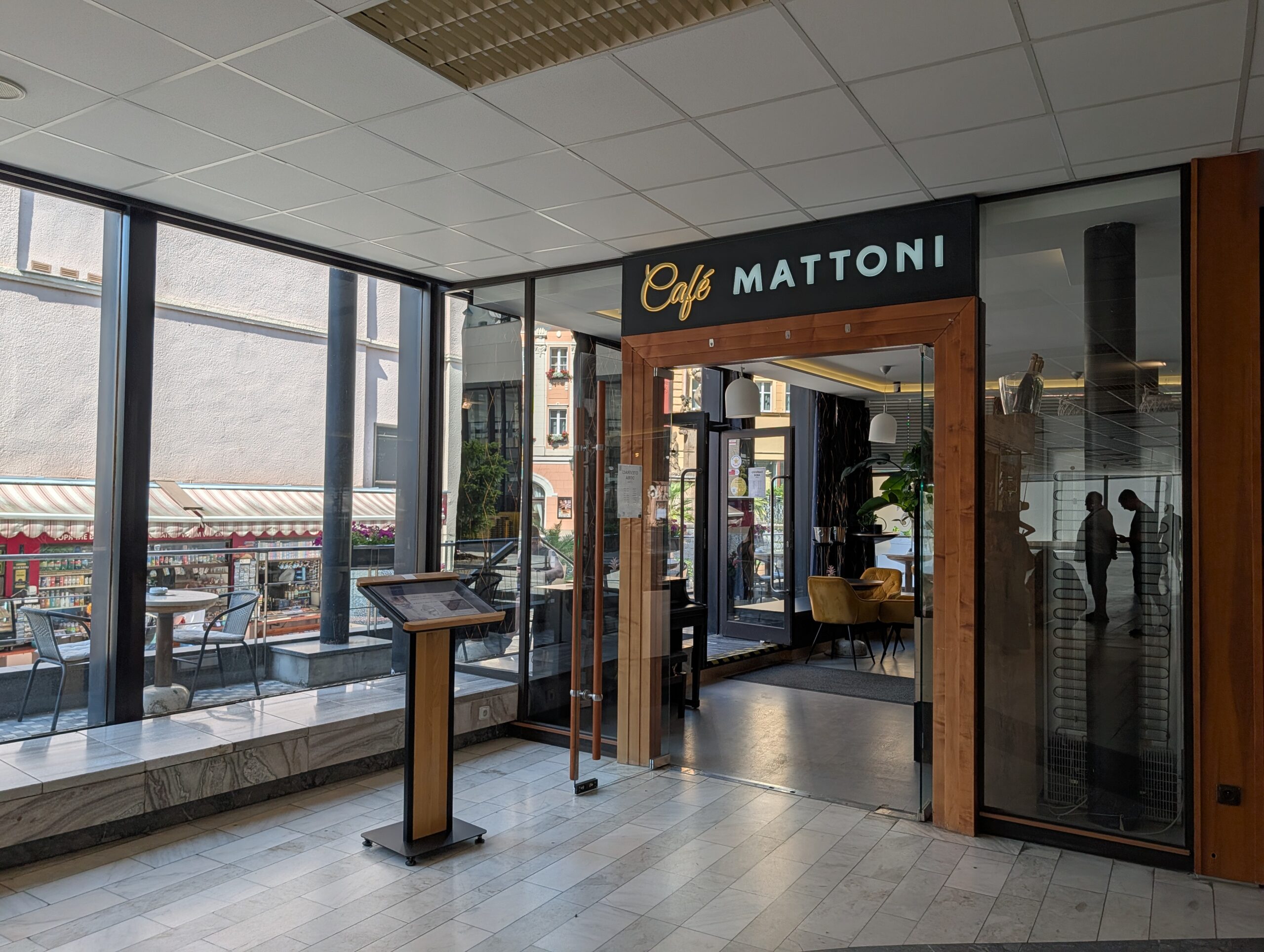 Eingang des Café Mattoni in Karlsbad mit Glasfront, Holzrahmen und Blick auf den Innenraum.