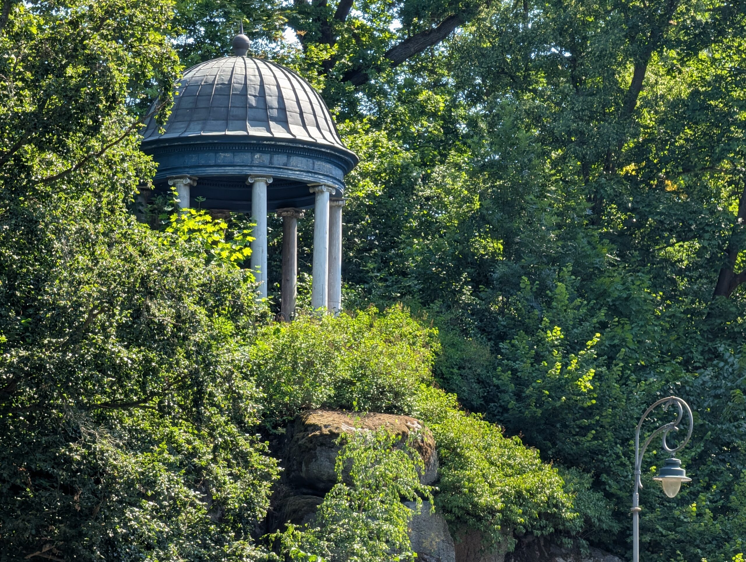 Dorotheen-Pavillon mit Kuppeldach und weißen Säulen zwischen Bäumen auf einem Felshang.