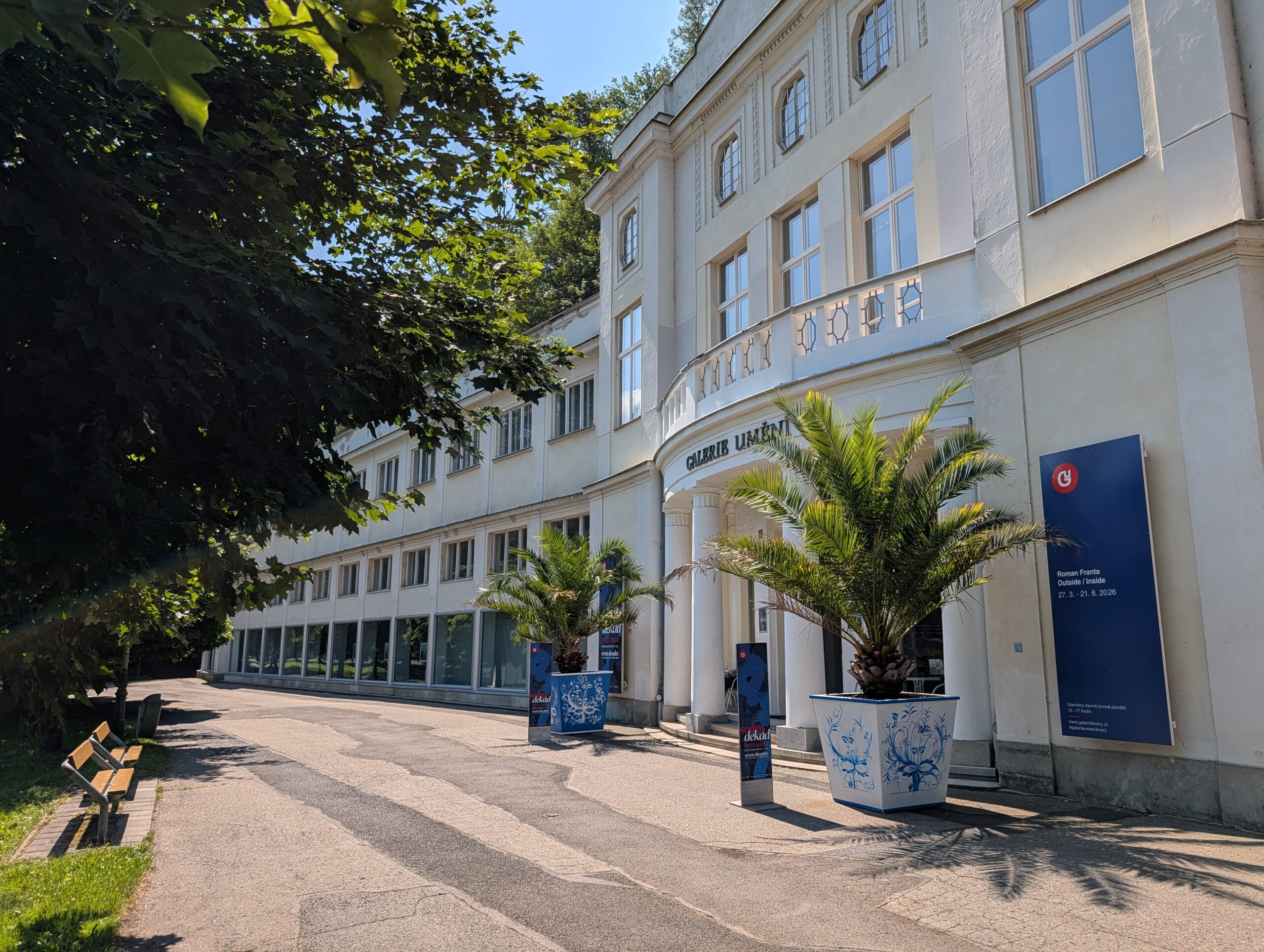 Helle Fassade der Kunstgalerie Karlsbad mit Eingang, Ausstellungstafeln und Palmen in Pflanzkübeln.