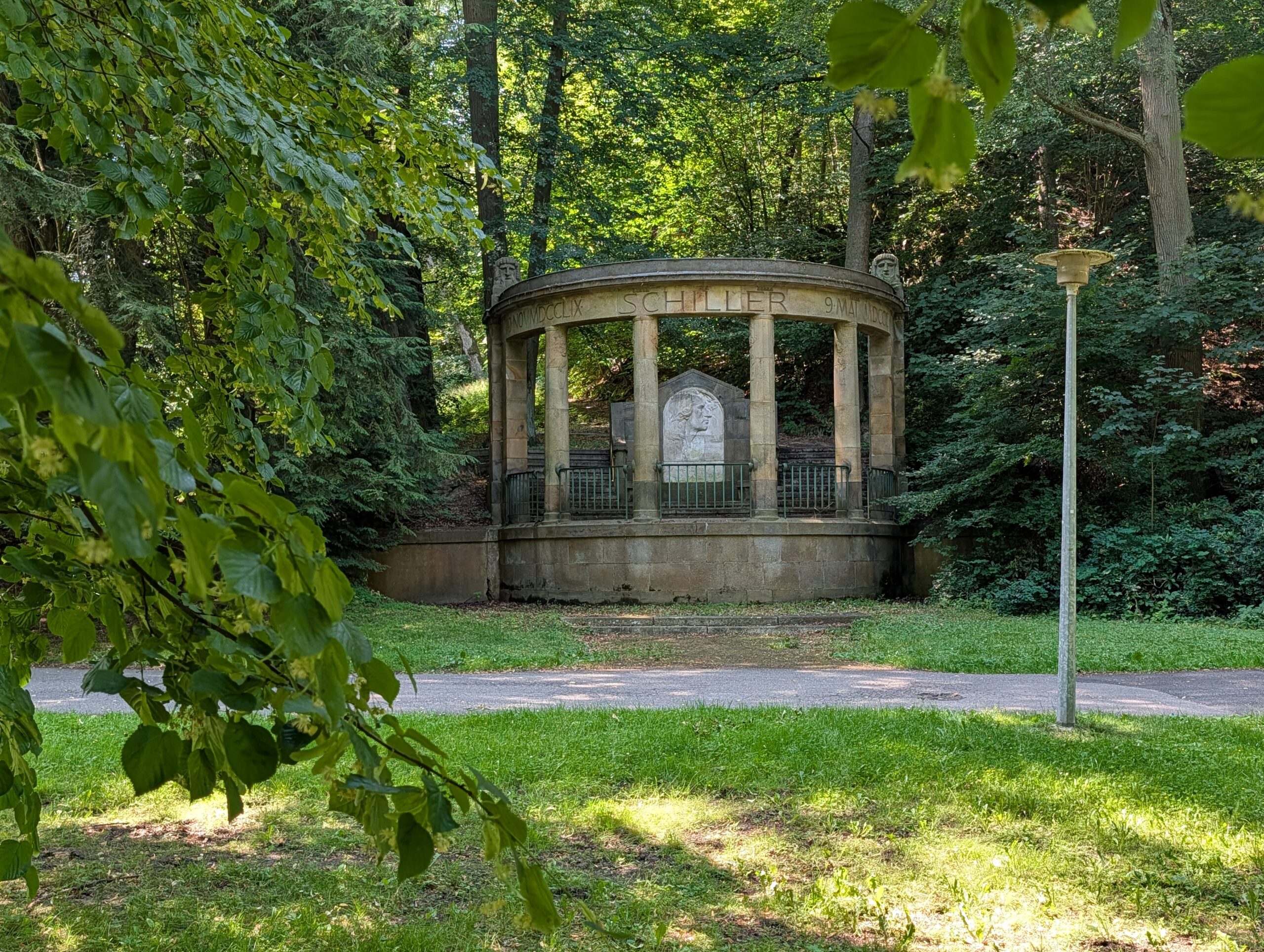 Friedrich-Schiller-Denkmal mit halbrunder Säulenanlage und Porträtrelief zwischen Bäumen und Sträuchern.