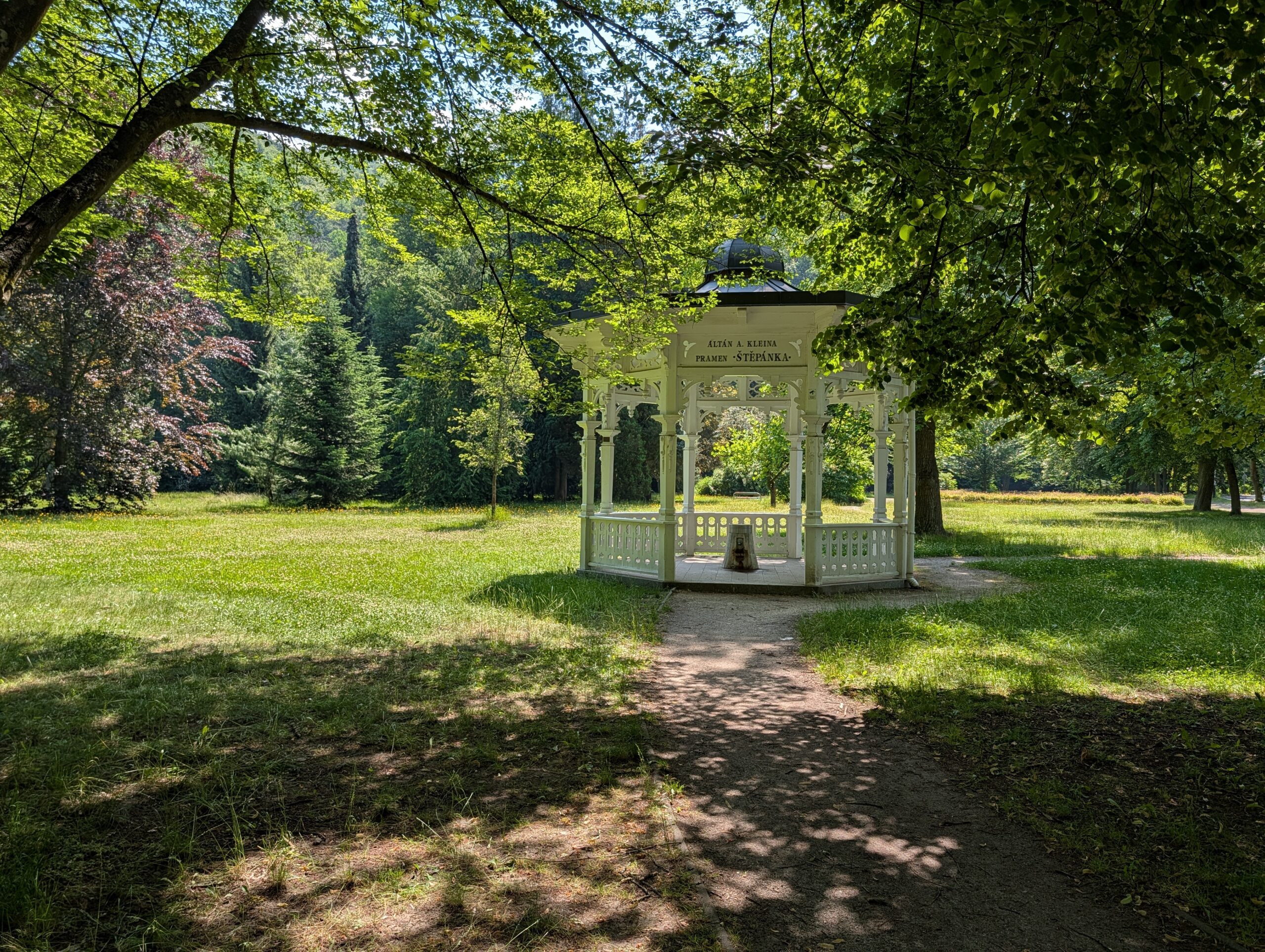 Weißer Holzpavillon der Stephanie-Quelle Nr. 14 auf einer grünen Parkwiese in Karlsbad.