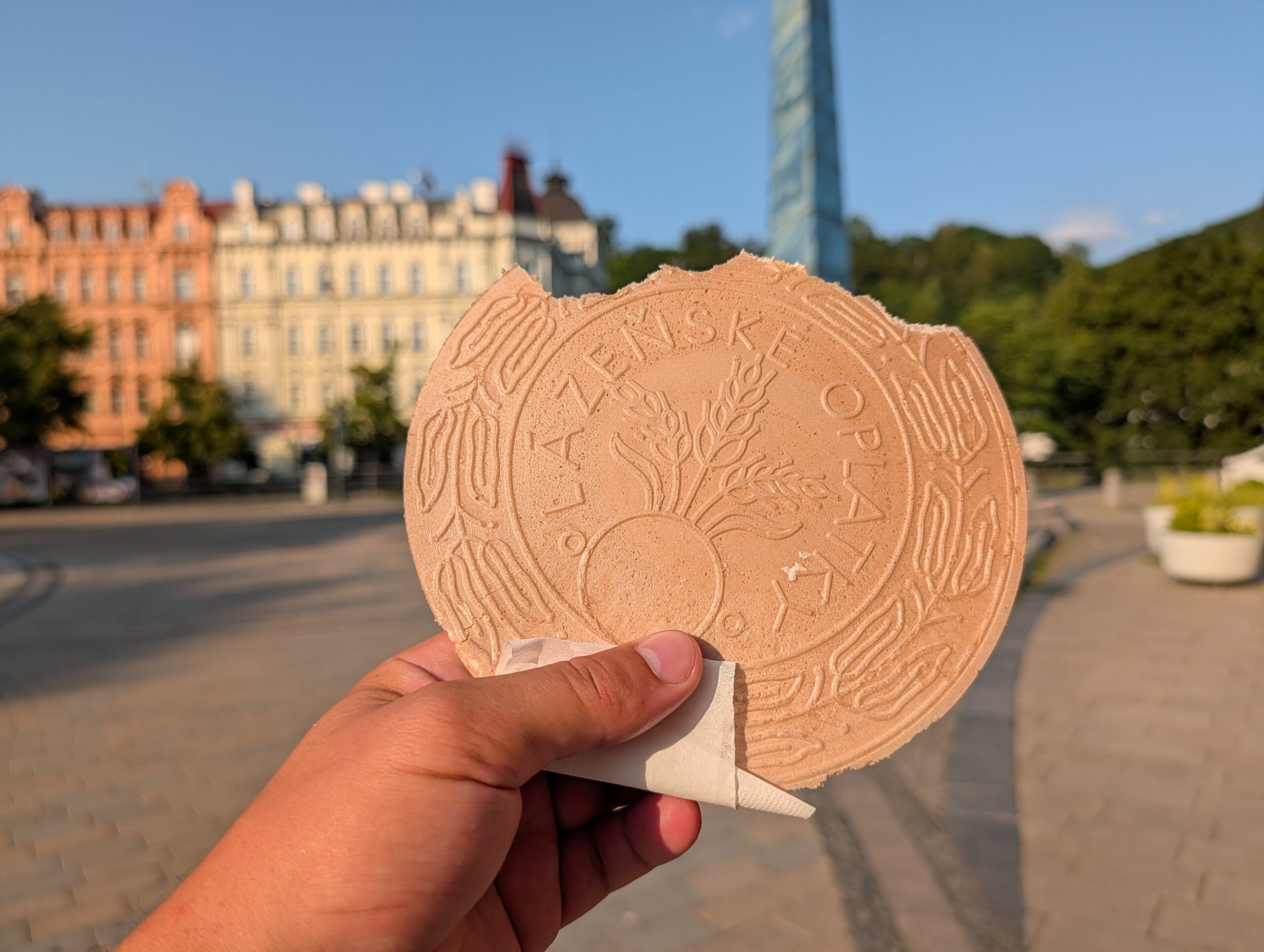 Hand hält eine Karlsbader Oblate (Karlovarská oplatka) vor dem Glasobelisk am T.-G.-Masaryk-Platz in Karlsbad.