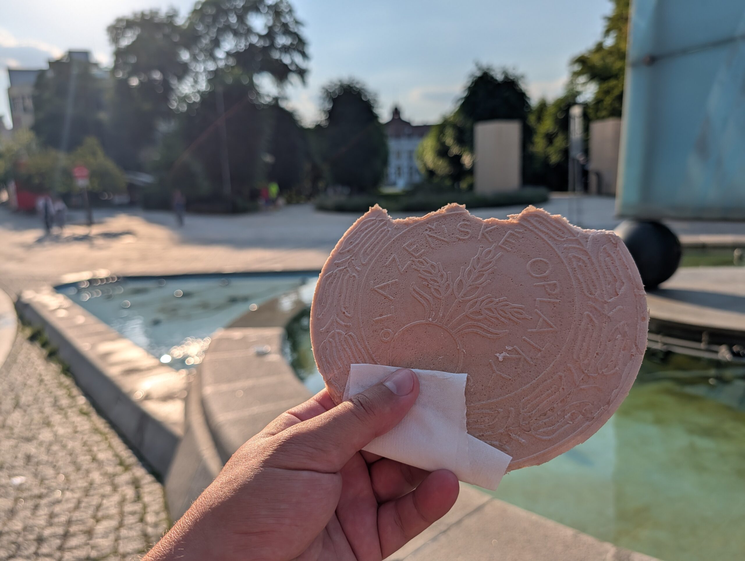 Hand hält eine Karlsbader Oblate (Karlovarská oplatka) vor einem Brunnen im Zentrum von Karlsbad.