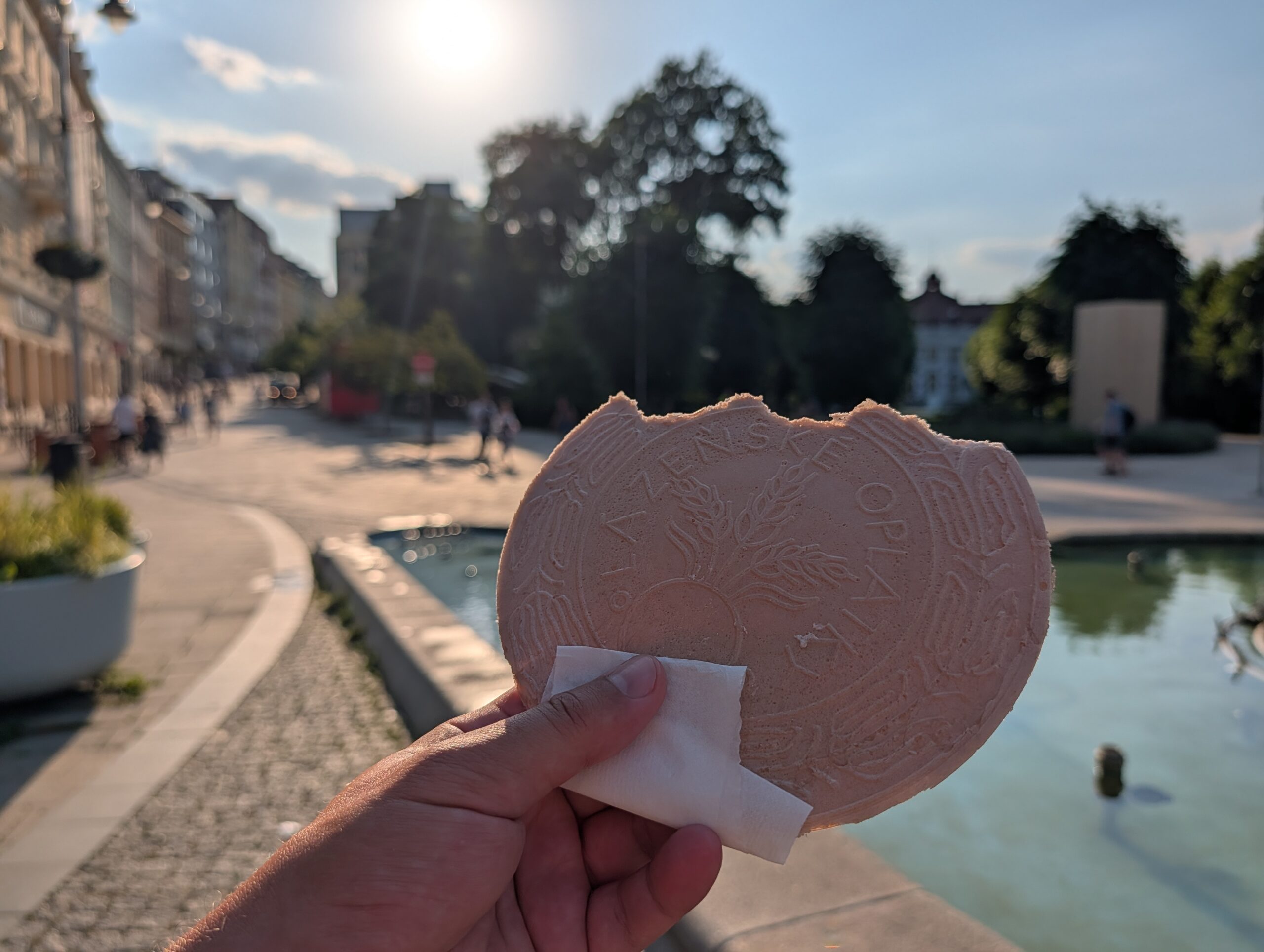 Hand hält eine Karlsbader Oblate (Karlovarská oplatka) vor einem Brunnen im Zentrum von Karlsbad.