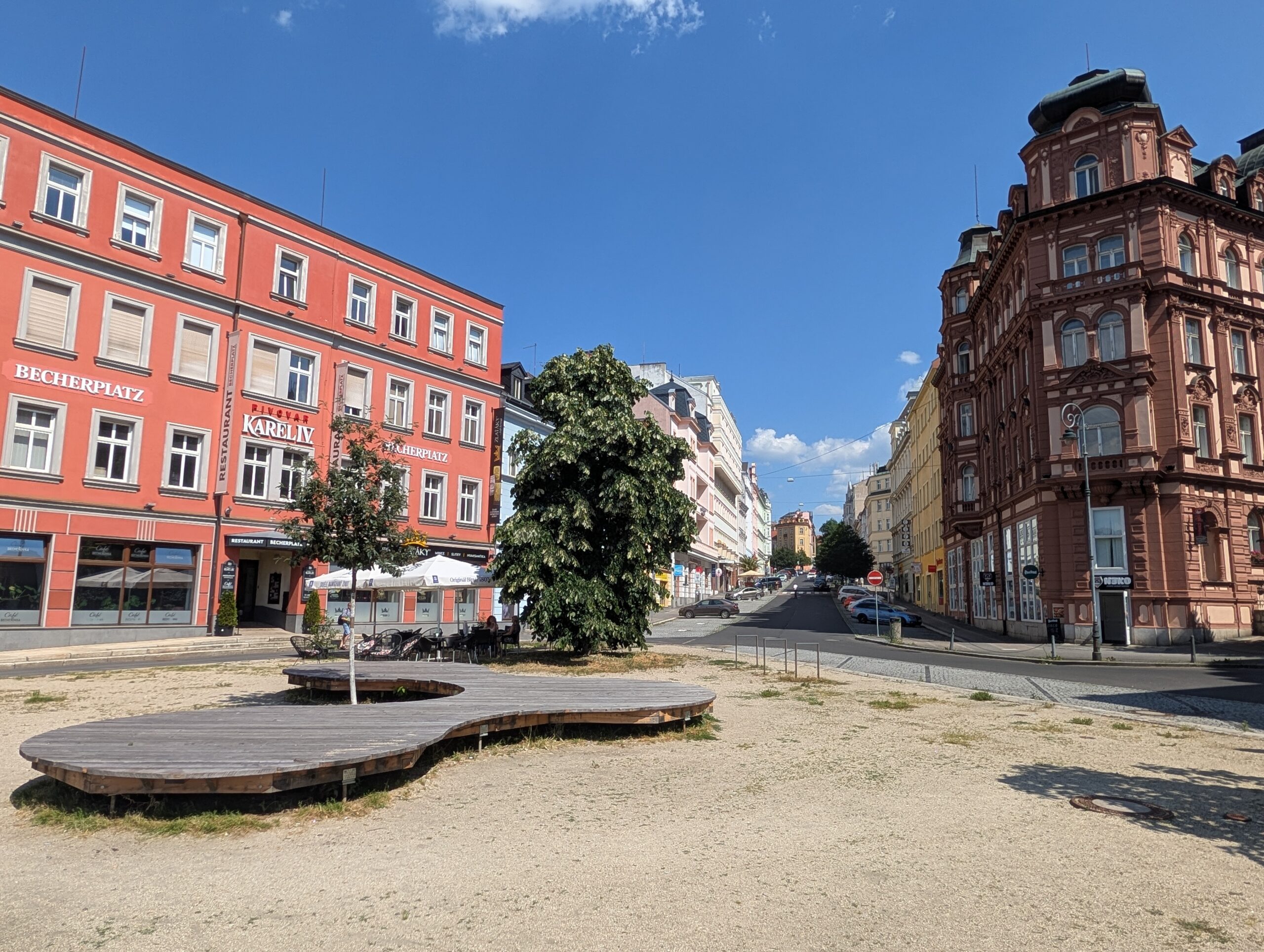 Offener Platz mit Sitzflächen, Bäumen, Straßen und mehrgeschossigen Gebäuden unter blauem Himmel.