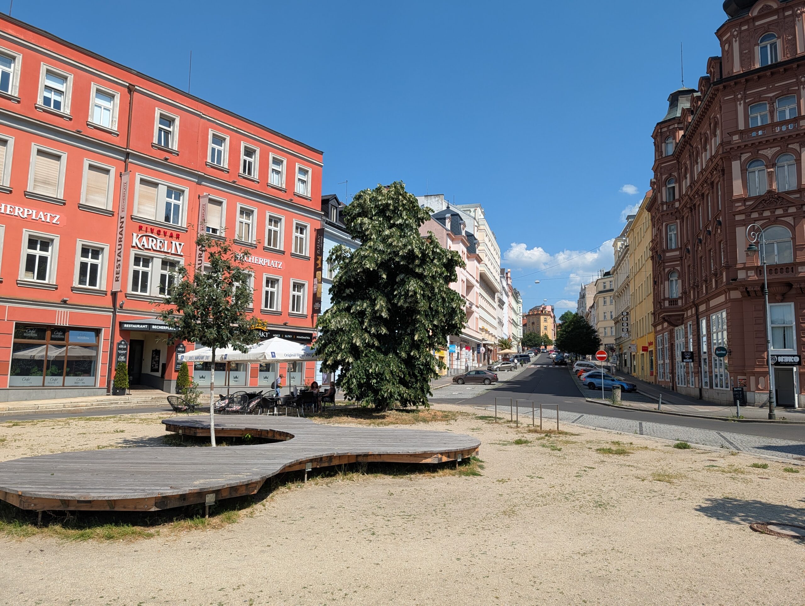 Offener Platz mit Holzsitzflächen, Bäumen, mehrgeschossigen Gebäuden und angrenzender Straße.