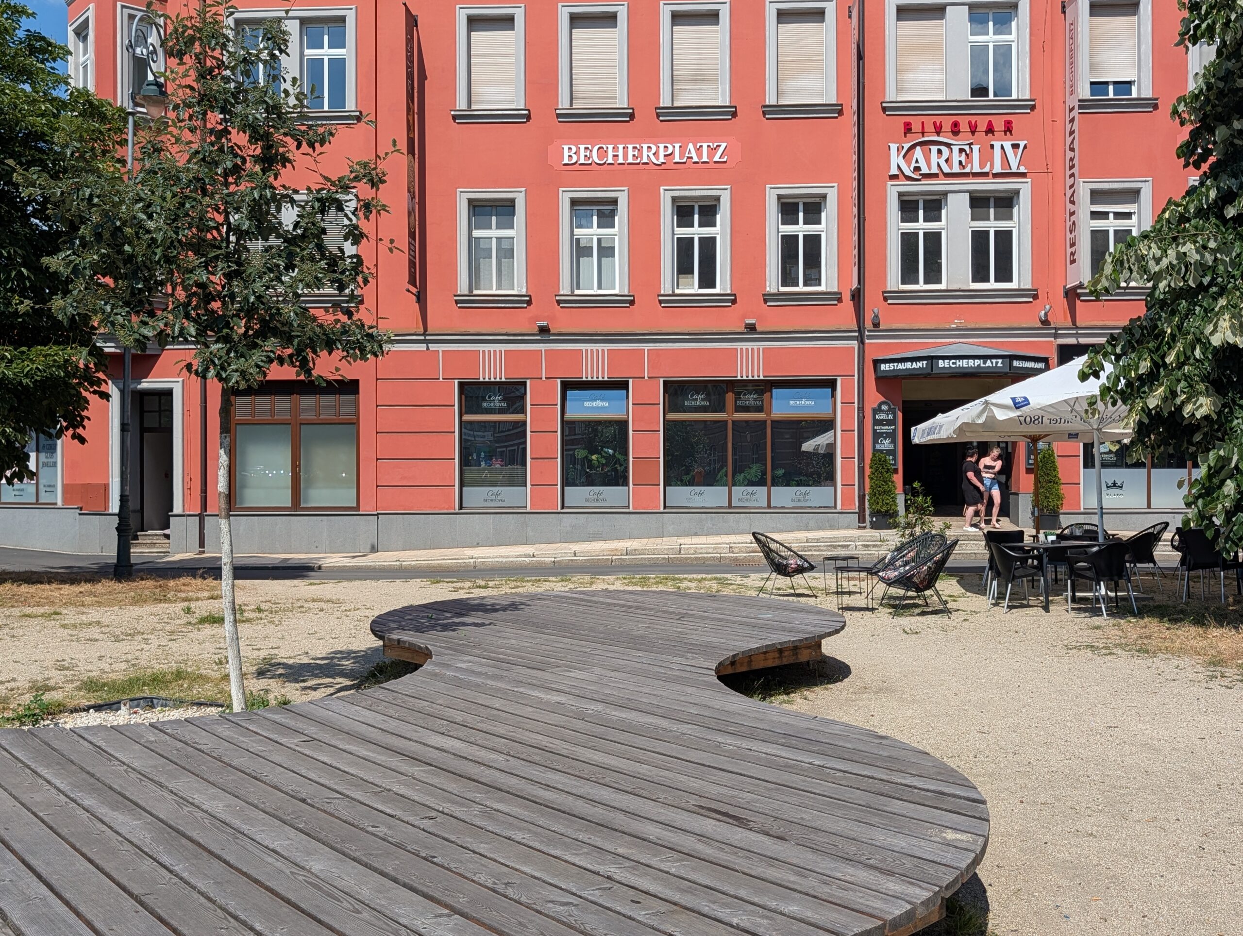 Holzsitzfläche auf einem Platz vor einem Gebäude mit Außengastronomie und der Beschriftung „BECHERPLATZ“ sowie „PIVOVAR KAREL IV“.