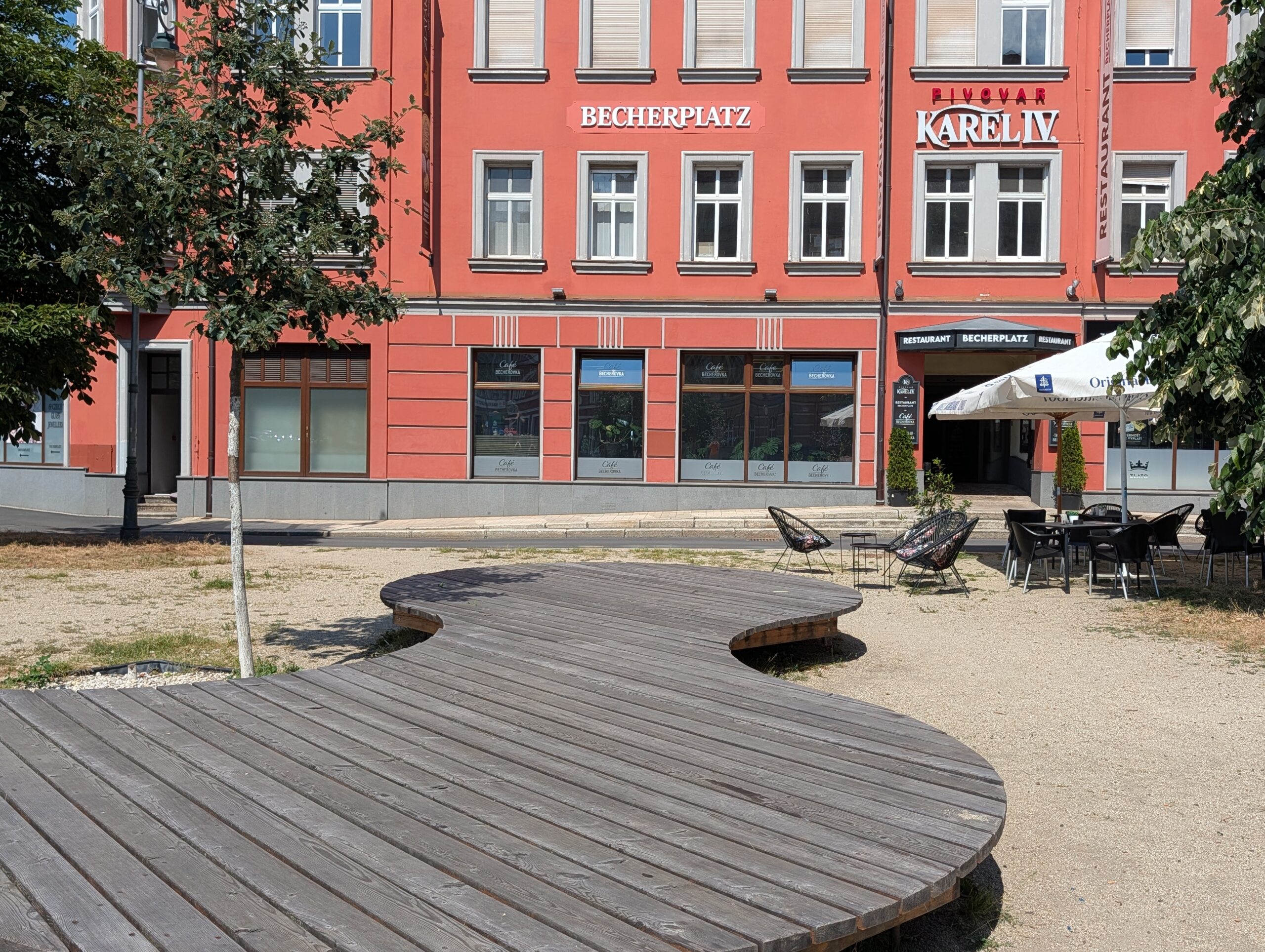 Holzsitzfläche auf einem Platz vor einem Gebäude mit Außengastronomie und der Beschriftung „BECHERPLATZ“ sowie „PIVOVAR KAREL IV“.