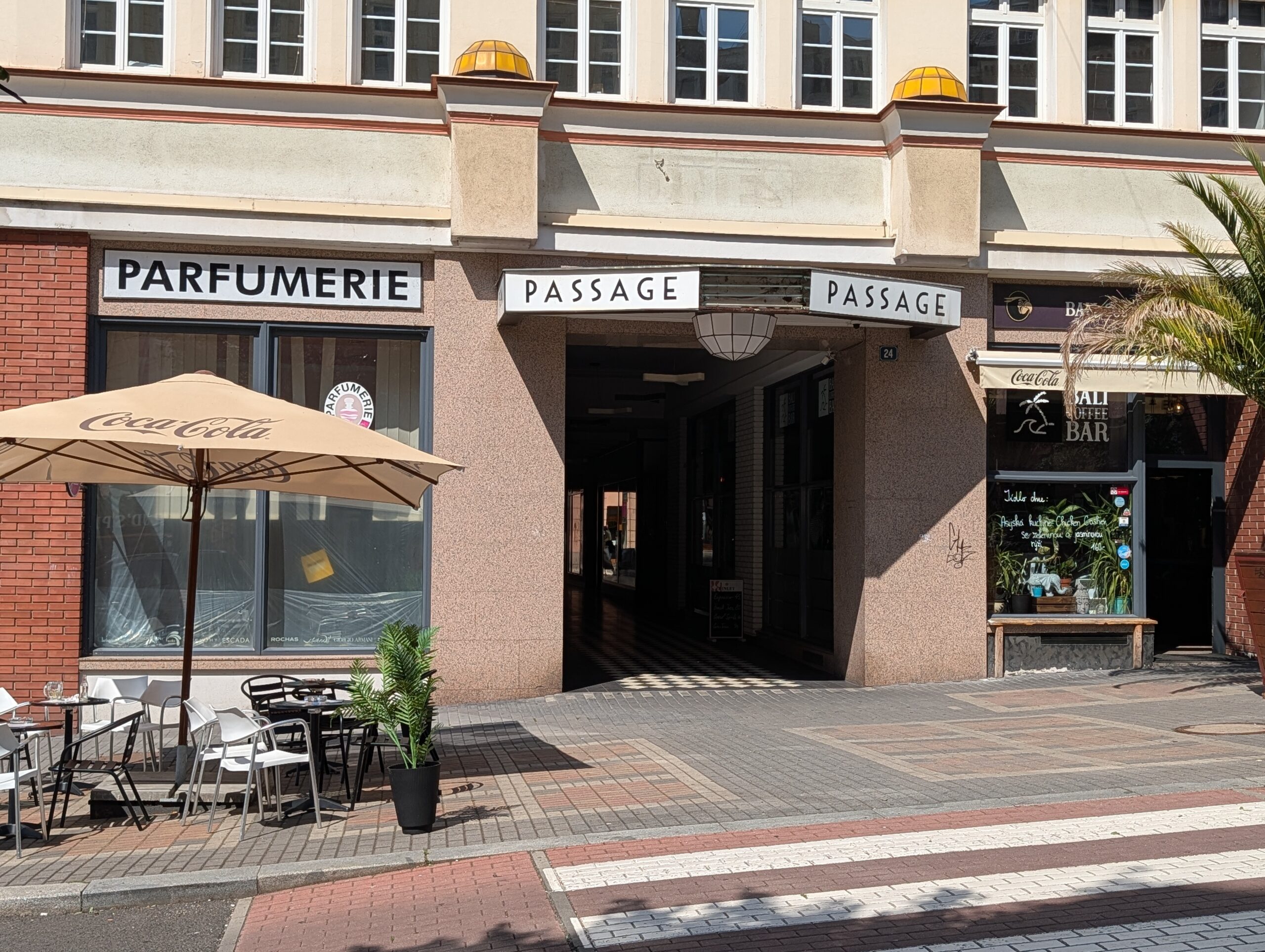Eingang einer überdachten Passage mit Geschäften, Schaufenstern, Außensitzplätzen und Fußgängerüberweg.