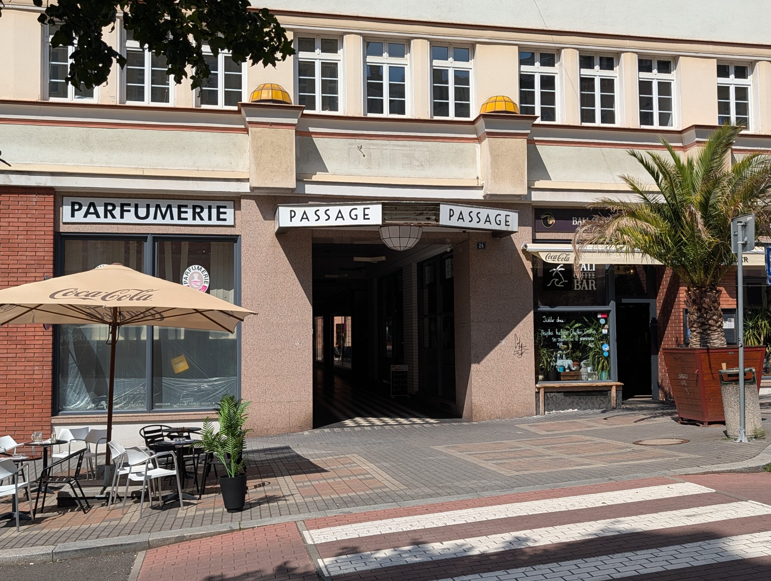 Eingang einer überdachten Passage mit Geschäften, Schaufenstern, Außensitzplätzen und Fußgängerüberweg.