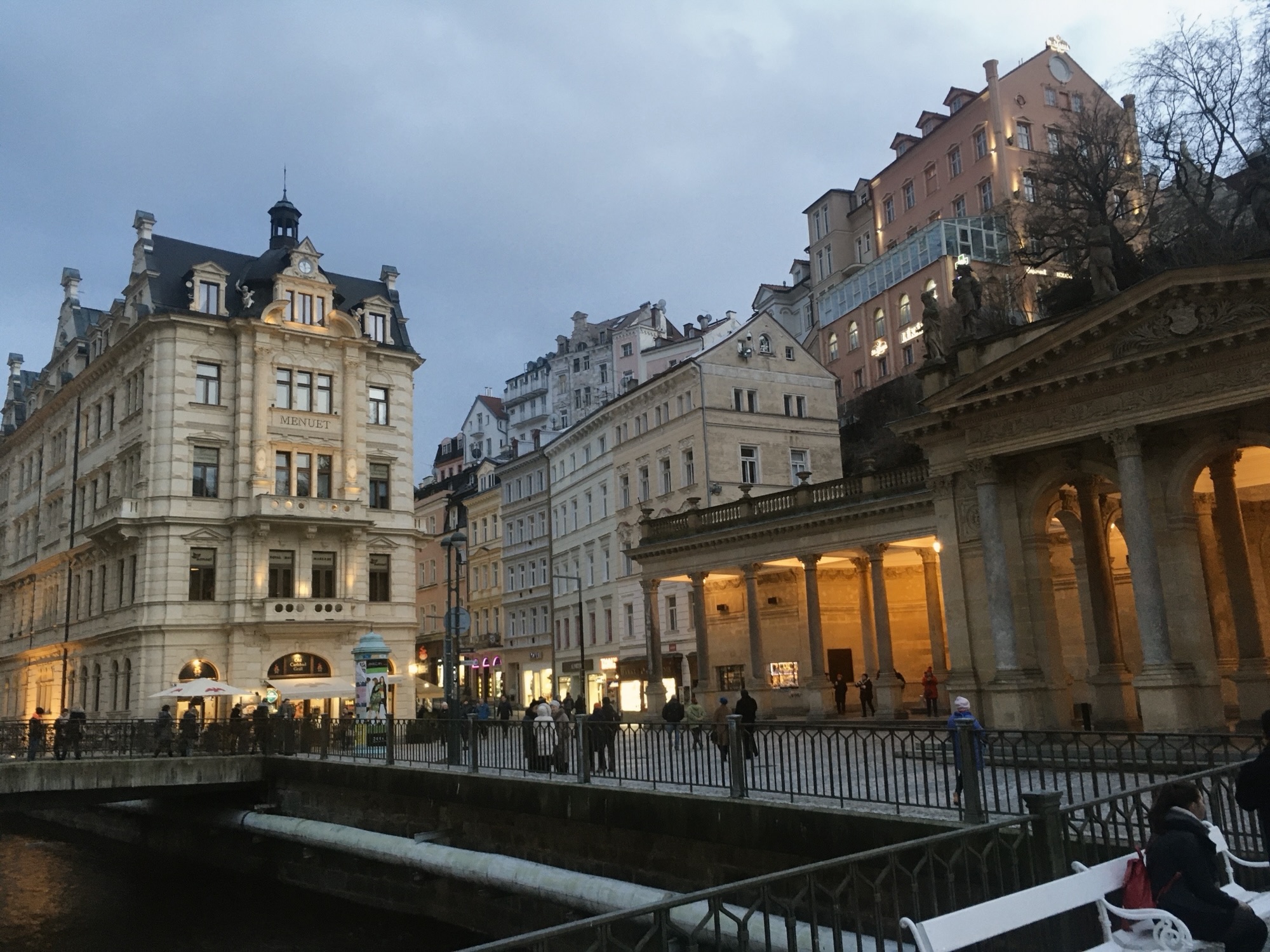 Beleuchtete Mühlbrunnkolonnade mit historischen Häusern, Brücke und Teplá am Abend